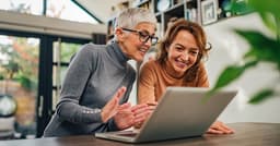 Two women are smiling and engaging with a laptop, sharing a joyful moment in a cozy indoor setting